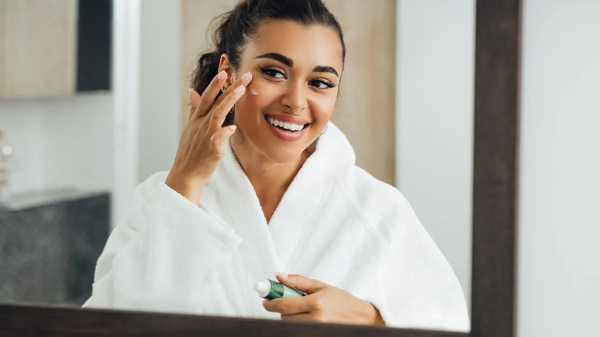 Middle east woman applying cream in bathroom mirror. Young smiling female putting moisturizer on her face. Copyright: xxArtemxVarnitsinx