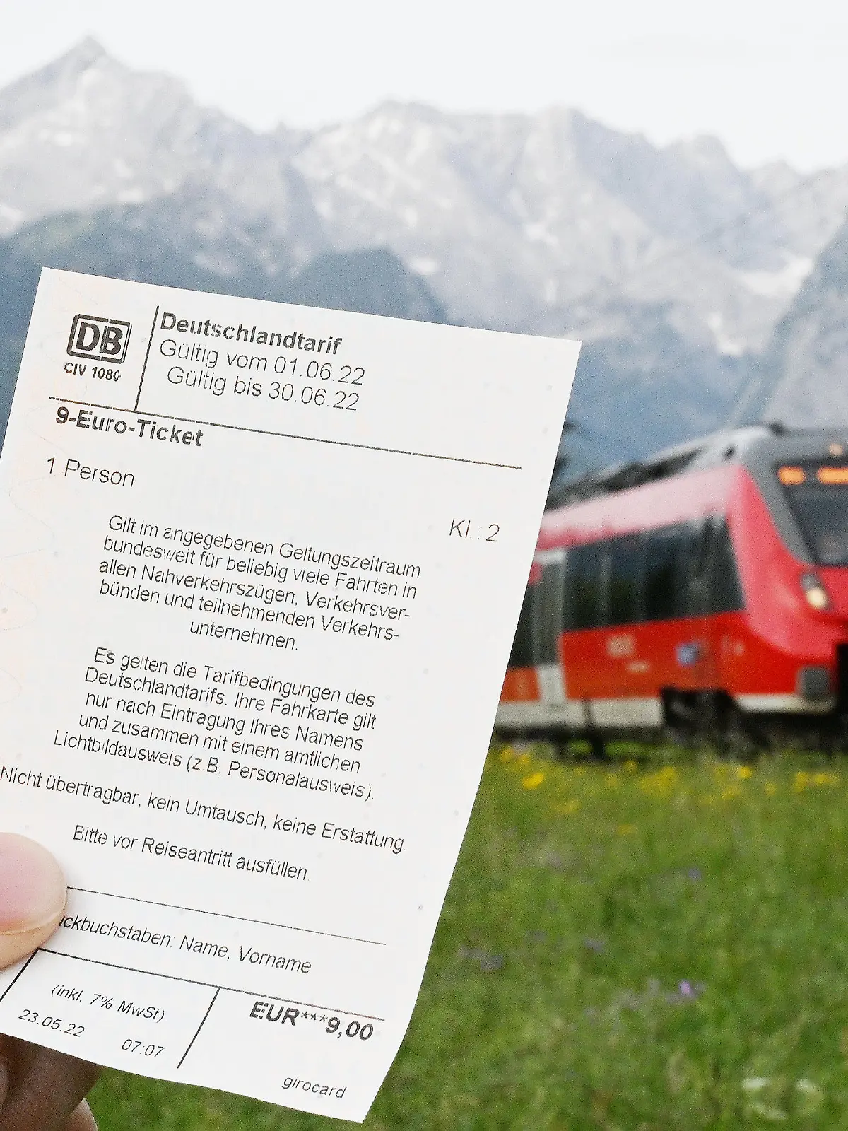 ARCHIV - 23.05.2022, Bayern, Garmisch-Partenkirchen: Ein Mann hält ein 9-Euro-Ticket in der Hand, im Hintergrund fährt ein Zug der Deutschen Bahn vor der Bergkulisse des Wettersteingebirges vorbei (gestelltes Foto). In Zügen auf dem Weg in die Alpen könnte es am langen Wochenende in Bayern wegen des günstigen Angebots voller werden. (zu dpa: «Ausflugsorte erwarten an Pfingsten wegen 9-Euro-Ticket mehr Andrang») Foto: Angelika Warmuth/dpa +++ dpa-Bildfunk +++