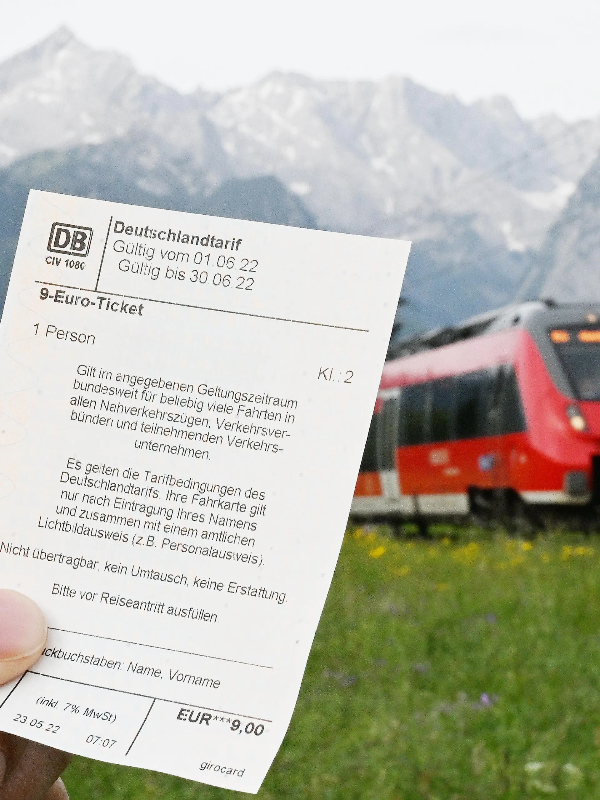 ARCHIV - 23.05.2022, Bayern, Garmisch-Partenkirchen: Ein Mann hält ein 9-Euro-Ticket in der Hand, im Hintergrund fährt ein Zug der Deutschen Bahn vor der Bergkulisse des Wettersteingebirges vorbei (gestelltes Foto). In Zügen auf dem Weg in die Alpen könnte es am langen Wochenende in Bayern wegen des günstigen Angebots voller werden. (zu dpa: «Ausflugsorte erwarten an Pfingsten wegen 9-Euro-Ticket mehr Andrang») Foto: Angelika Warmuth/dpa +++ dpa-Bildfunk +++