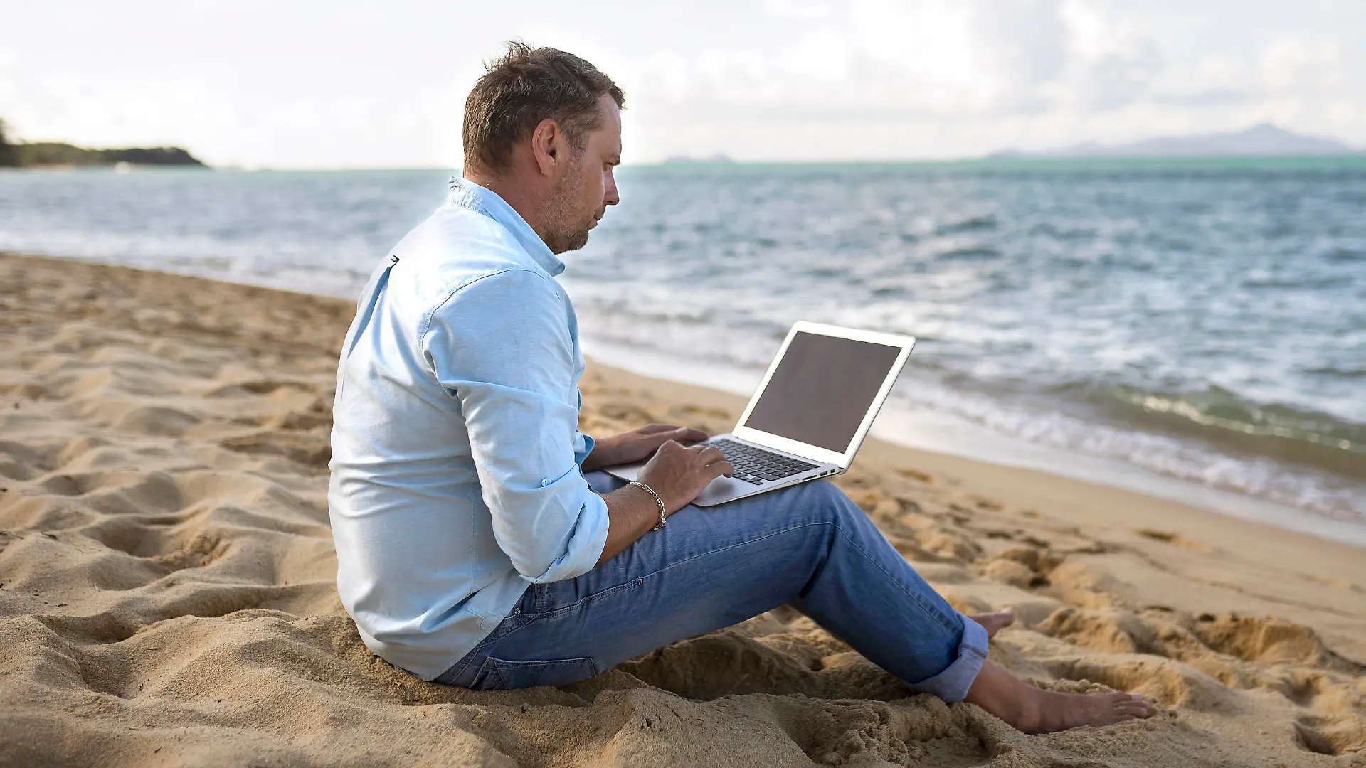 Caucasian man sitting on beach using laptop, Kaukasischer Mann sitzt am Strand mit Laptop || Modellfreigabe vorhanden