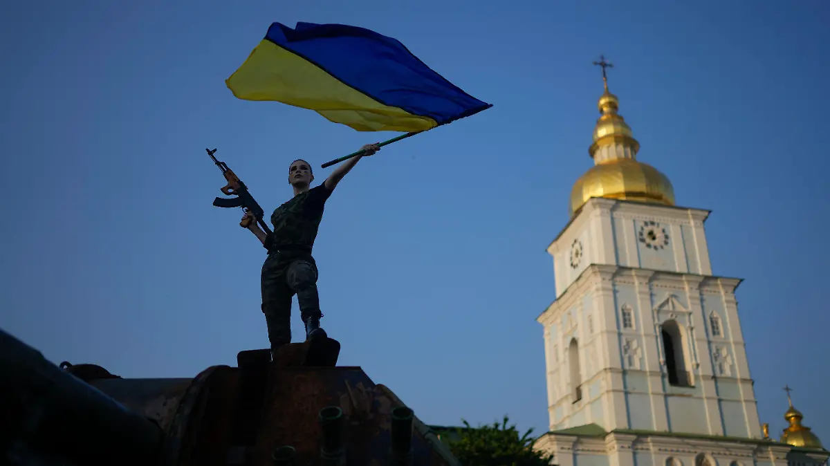 10.06.2022, Ukraine, Kiew: Eine Frau schwenkt die ukrainische Flagge auf einem zerstörten russischen Panzer. Die Sonne scheint, aber es herrschen Traurigkeit und grimmige Entschlossenheit. Der Sommer 2022 ist für die ukrainische Hauptstadt Kiew bitter, denn an den Fronten im Osten und Süden tobt der Krieg. In gut dreieinhalb Monaten des russischen Angriffskrieges sind nach Regierungsangaben etwa 10.000 ukrainische Soldaten getötet worden. Foto: Natacha Pisarenko/AP/dpa +++ dpa-Bildfunk +++