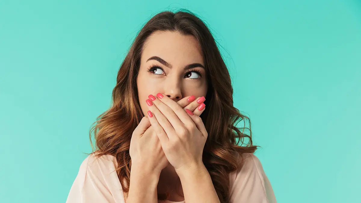 Portrait of a lovely young girl in dress standing with mouth covered and looking away at copy space isolated over blue background
