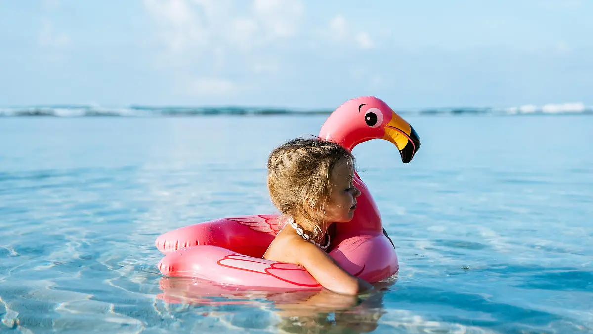 4 years old little girl, wearing necklace made of seashells, swimming in a tropical ocean on inflatable pink flamingo. Blue sky with few fluffy clouds on the background.