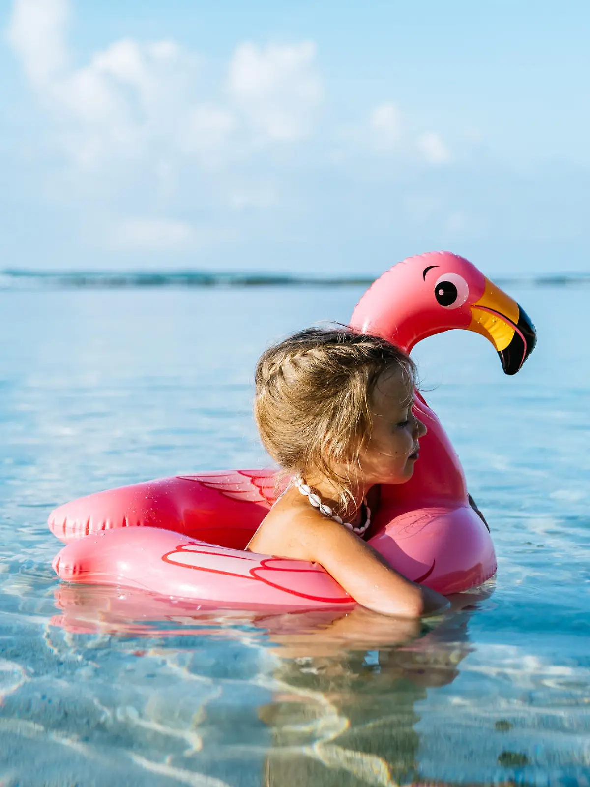 4 years old little girl, wearing necklace made of seashells, swimming in a tropical ocean on inflatable pink flamingo. Blue sky with few fluffy clouds on the background.