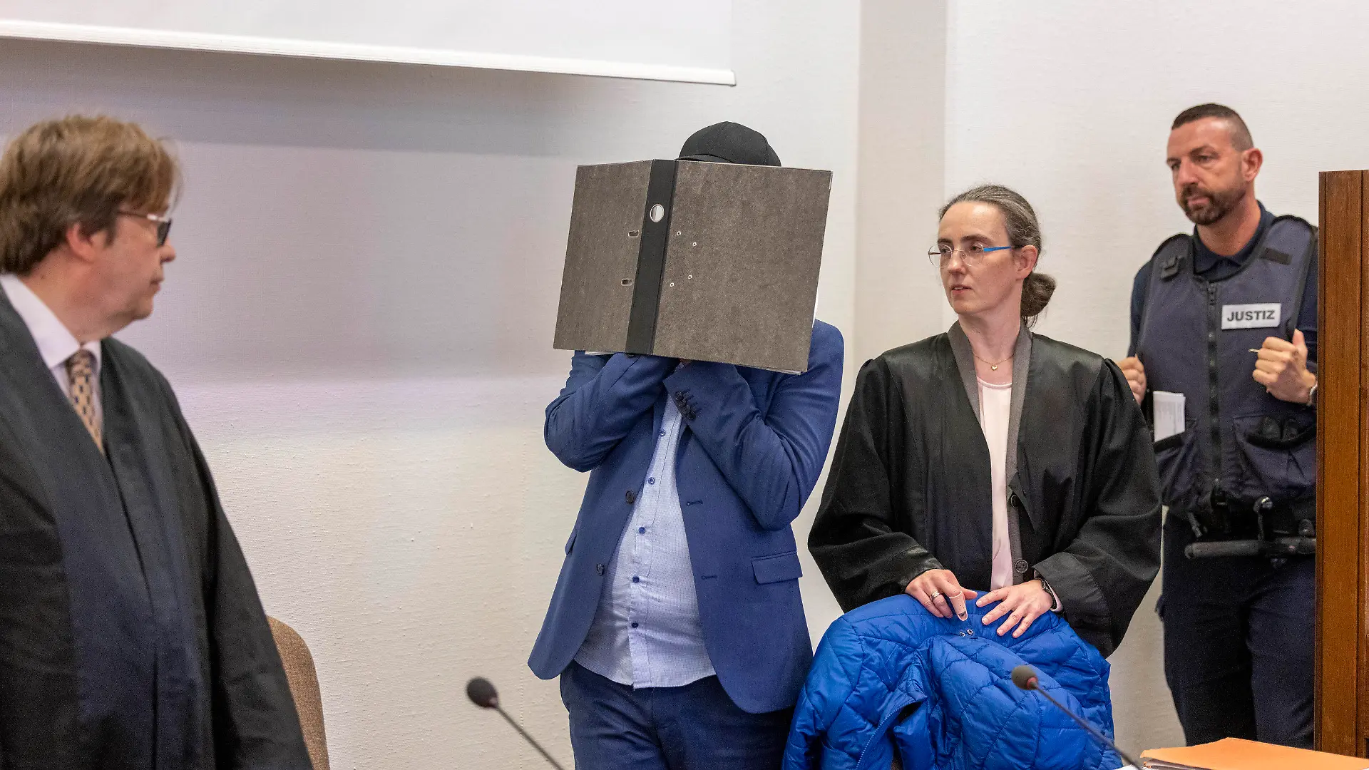 29.08.2023, Nordrhein-Westfalen, Köln: Der Angeklagte kommt vor der Verkündung des Urteils im Prozess wegen sexuellen Missbrauchs an Kindern in den Gerichtssaal des Kölner Landgerichts, während er sein Gesicht hinter einem Aktenordner verbirgt. Seine beiden Rechtsanwälte Jürgen Schüttler (l) und Sybille Weber (r) erwarten den Angeklagten im Gerichtssaal. In Köln wird am Dienstag (11.00 Uhr) das Urteil in einem Missbrauchsprozess gegen einen ehemaligen Babysitter erwartet. Der 34-Jährige soll über Jahre als Babysitter und flexible Aushilfskraft in Kindertagesstätten in Köln gearbeitet und dabei Kindern sexuelle Gewalt angetan haben. Foto: Thomas Banneyer/dpa +++ dpa-Bildfunk +++