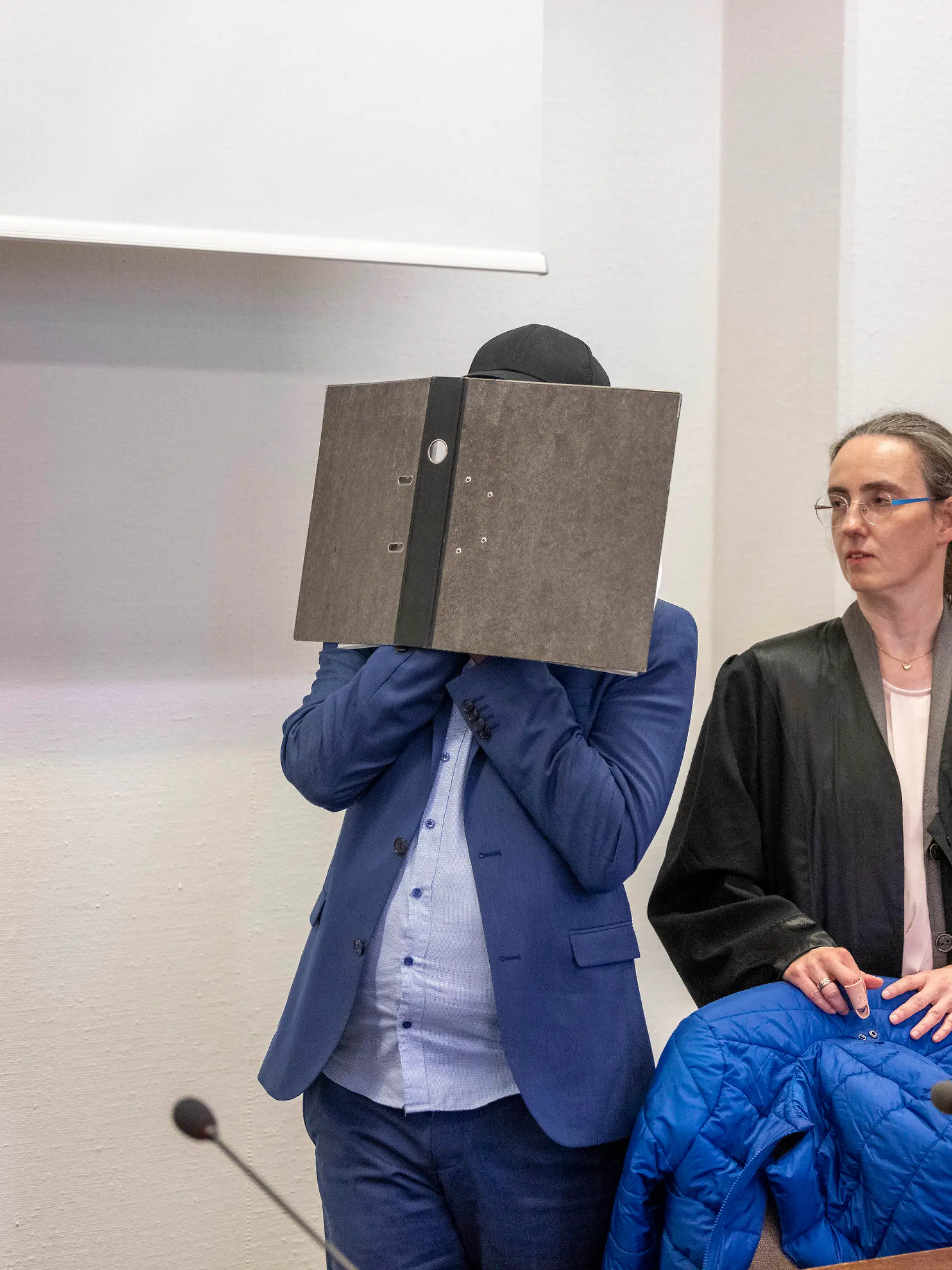 29.08.2023, Nordrhein-Westfalen, Köln: Der Angeklagte kommt vor der Verkündung des Urteils im Prozess wegen sexuellen Missbrauchs an Kindern in den Gerichtssaal des Kölner Landgerichts, während er sein Gesicht hinter einem Aktenordner verbirgt. Seine beiden Rechtsanwälte Jürgen Schüttler (l) und Sybille Weber (r) erwarten den Angeklagten im Gerichtssaal. In Köln wird am Dienstag (11.00 Uhr) das Urteil in einem Missbrauchsprozess gegen einen ehemaligen Babysitter erwartet. Der 34-Jährige soll über Jahre als Babysitter und flexible Aushilfskraft in Kindertagesstätten in Köln gearbeitet und dabei Kindern sexuelle Gewalt angetan haben. Foto: Thomas Banneyer/dpa +++ dpa-Bildfunk +++