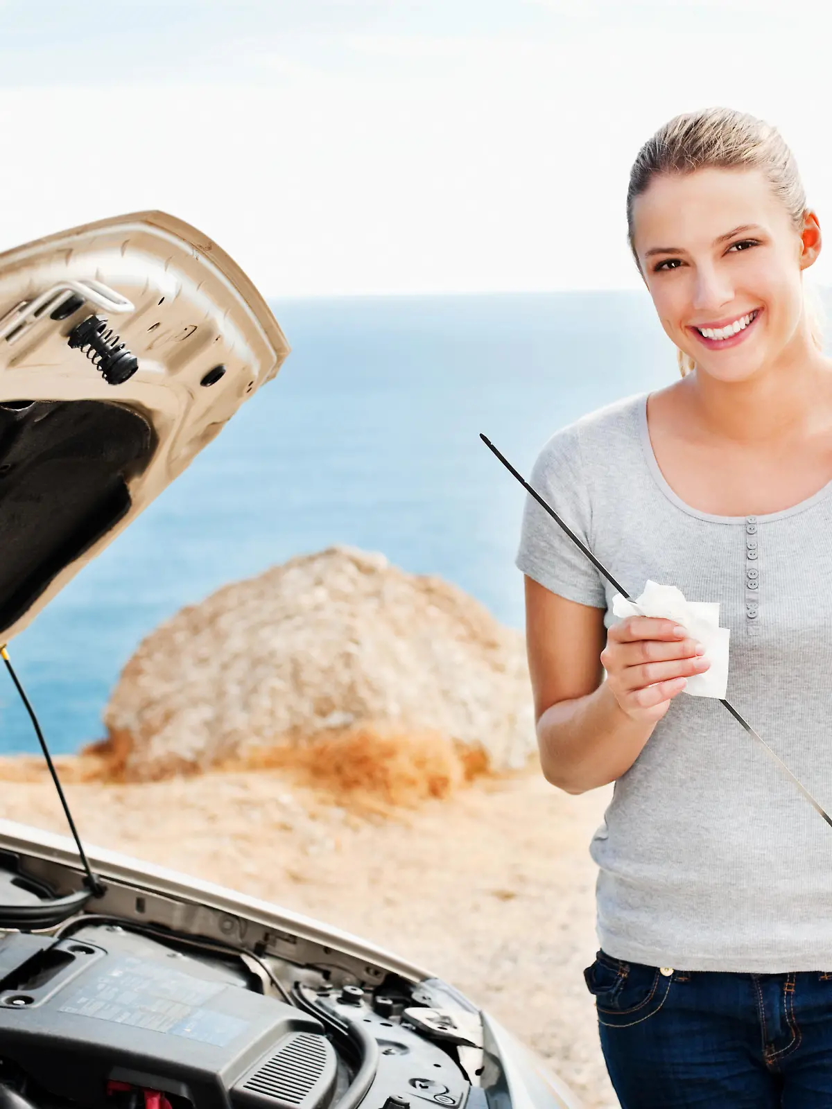 A young woman smiles towards the camera while checking the oil in her car.  Horizontal shot.