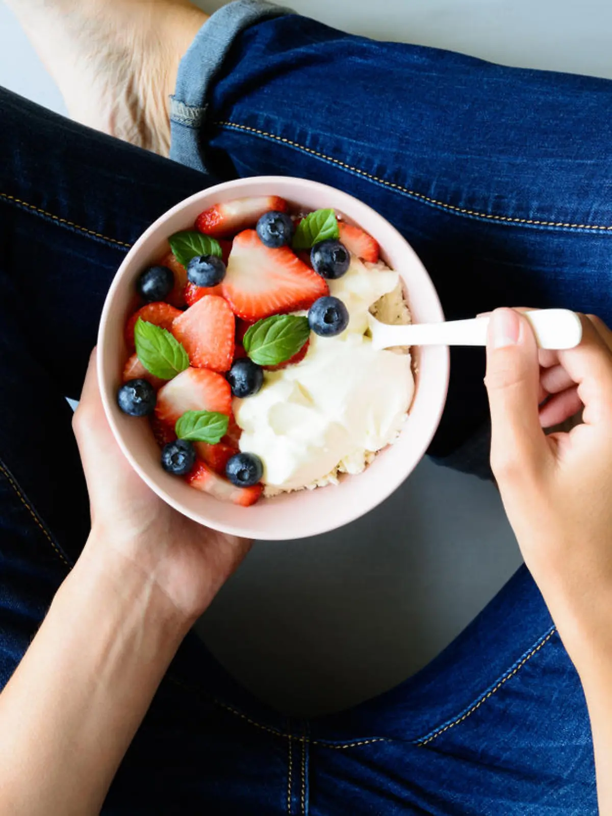 Woman hands holding bowl with cottage cheese, cream, strawberry, blueberry and mint leaves on gray background. Top view. Healthy lifestyle