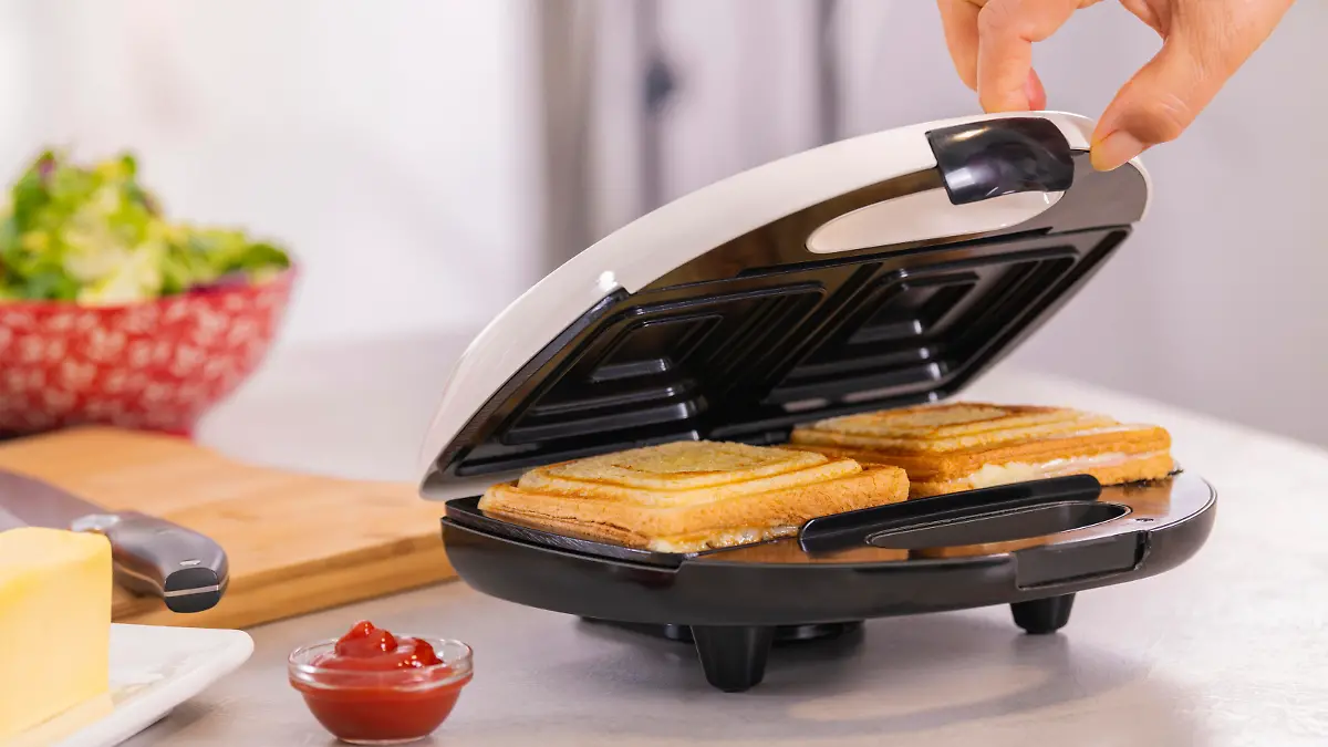 Female hand carefully opening the lid of a sandwich making machine with hot sandwiches. Selective focus and close up. Homemade food concept.