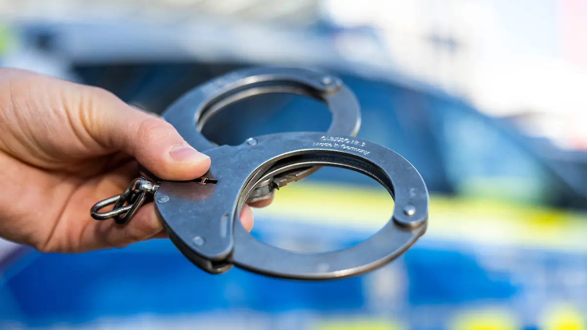 ARCHIV - 28.09.2021, Nordrhein-Westfalen, Gütersloh: Eine Hand hält Handschellen vor einen Streifenwagen der Polizei. Am 05.04.2022 wird in Berlin die Polizeiliche Kriminalstatistik 2021 vorgestellt. Foto: David Inderlied/dpa +++ dpa-Bildfunk +++
