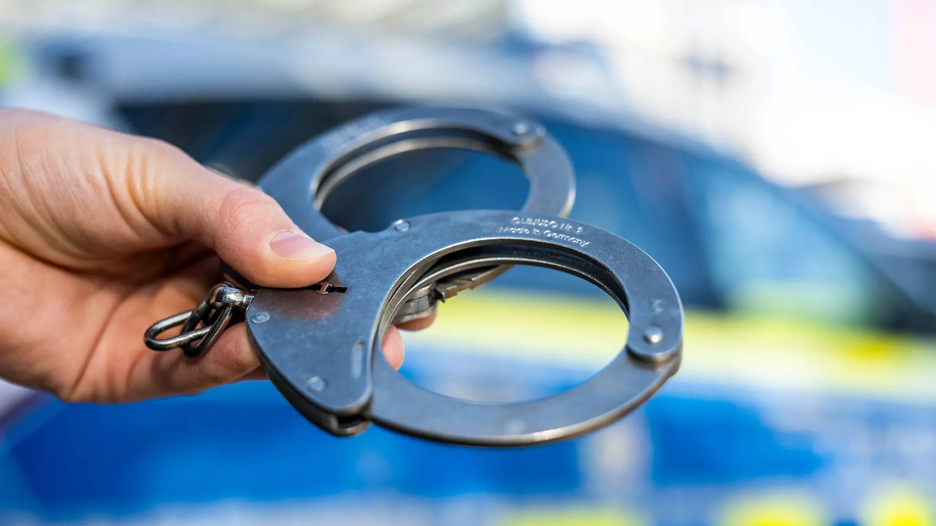 ARCHIV - 28.09.2021, Nordrhein-Westfalen, Gütersloh: Eine Hand hält Handschellen vor einen Streifenwagen der Polizei. Am 05.04.2022 wird in Berlin die Polizeiliche Kriminalstatistik 2021 vorgestellt. Foto: David Inderlied/dpa +++ dpa-Bildfunk +++