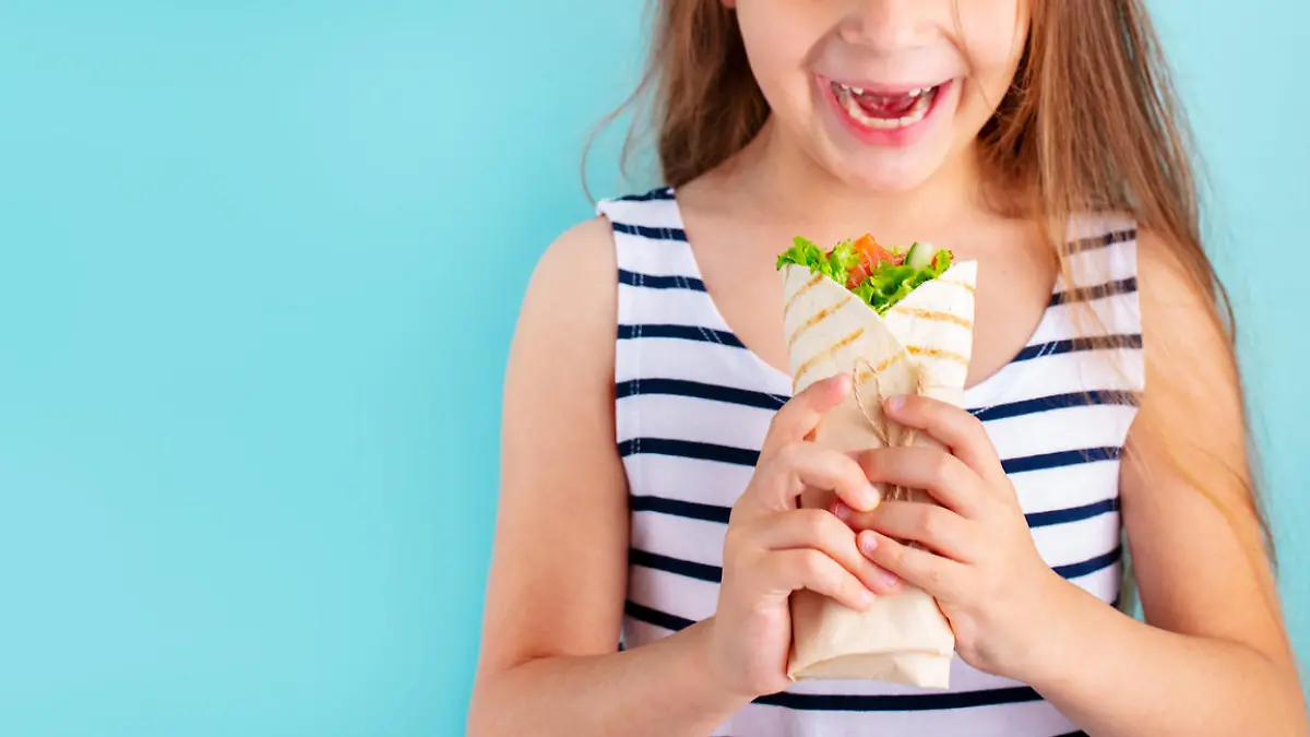 Smiling beautiful child girl eating wrap sandwich. Blue background. Copy space.