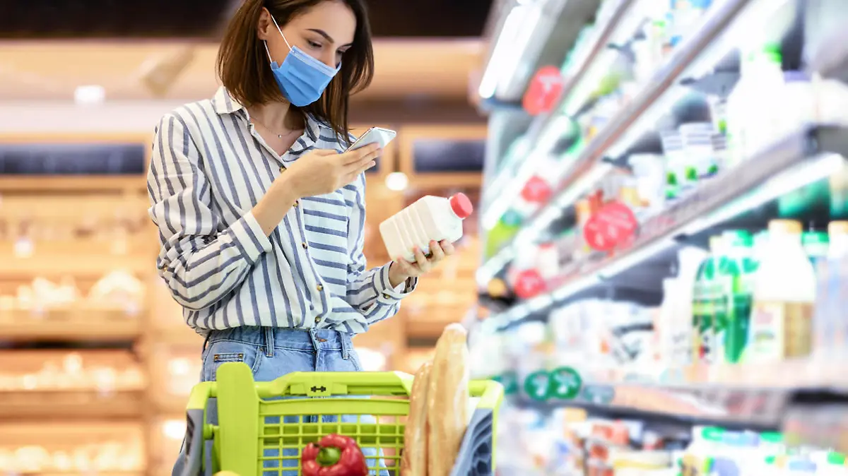 Young woman in disposable face mask taking dairy products from shelf in the supermarket, holding bottle and smartphone, scanning bar code on product through mobile phone, walking with trolley cart