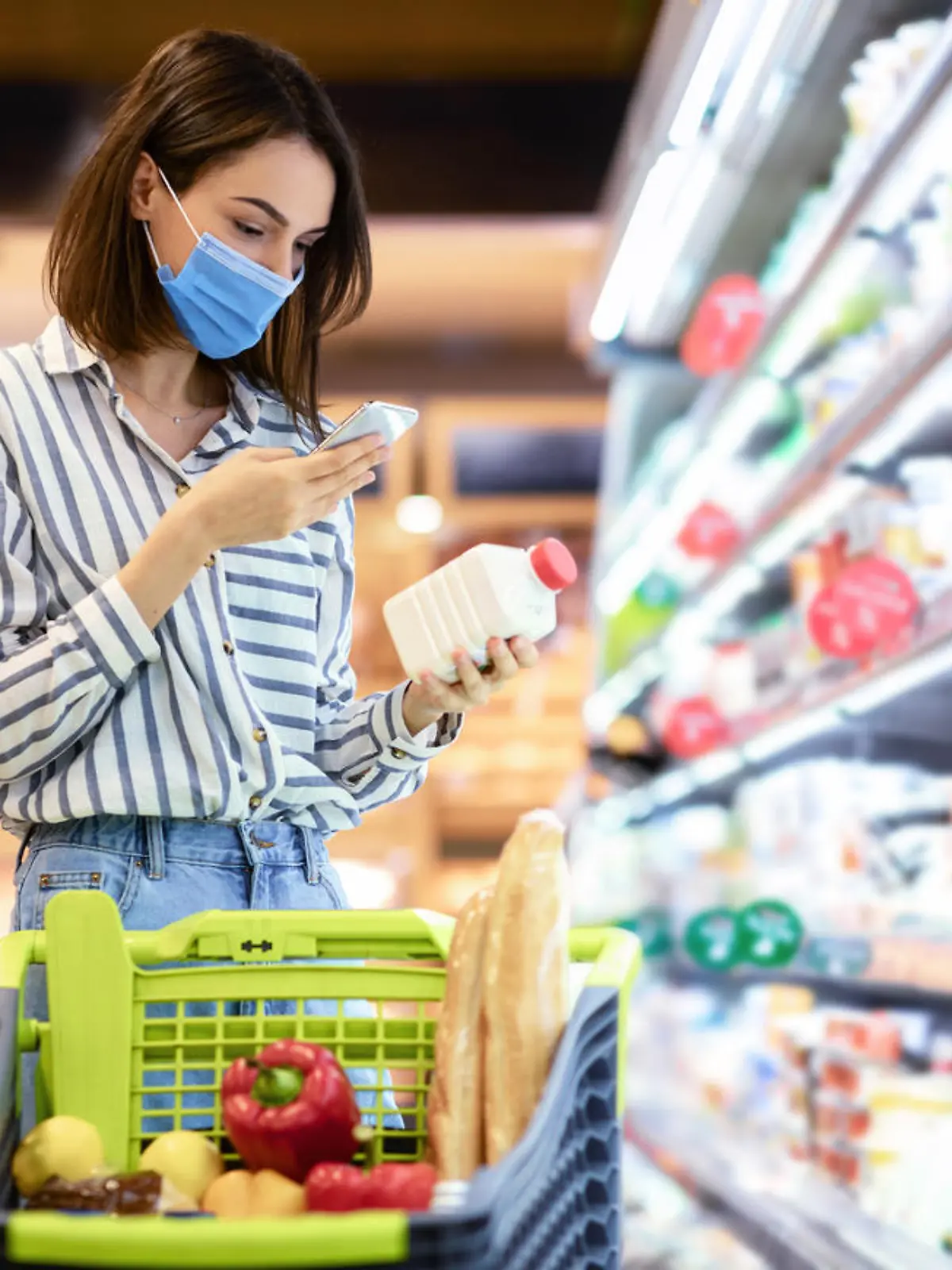 Young woman in disposable face mask taking dairy products from shelf in the supermarket, holding bottle and smartphone, scanning bar code on product through mobile phone, walking with trolley cart