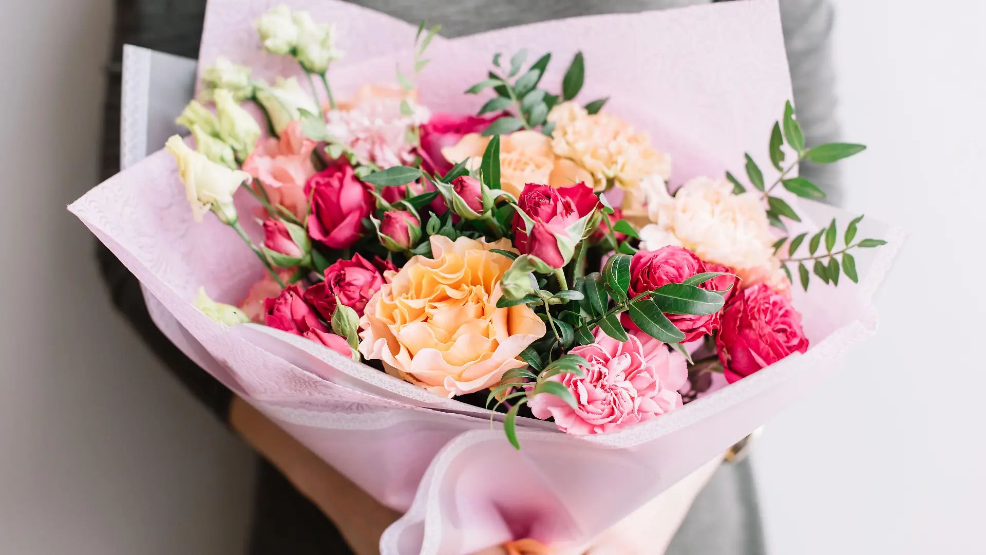 Very nice young woman holding a colourful fresh blossoming flower bouquet of different sorts of roses, carnations, eustoma, peonies on the grey wall background