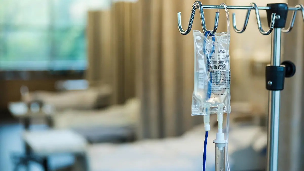 The focus of the photo is on the IV bag and stand in the foreground with empty hospital beds in a row in the background.