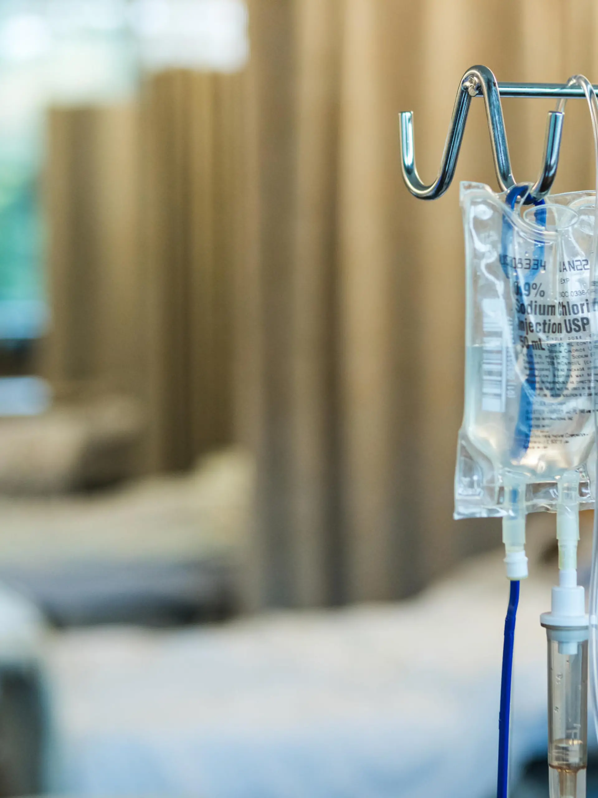 The focus of the photo is on the IV bag and stand in the foreground with empty hospital beds in a row in the background.