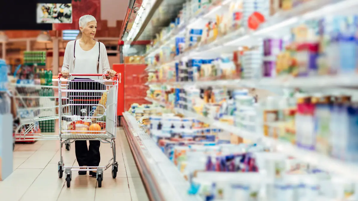 Mature smiling woman shopping in local supermarket. She is shopping groceries. Walking by refrigerator with dairy product and pushing shopping trolley.