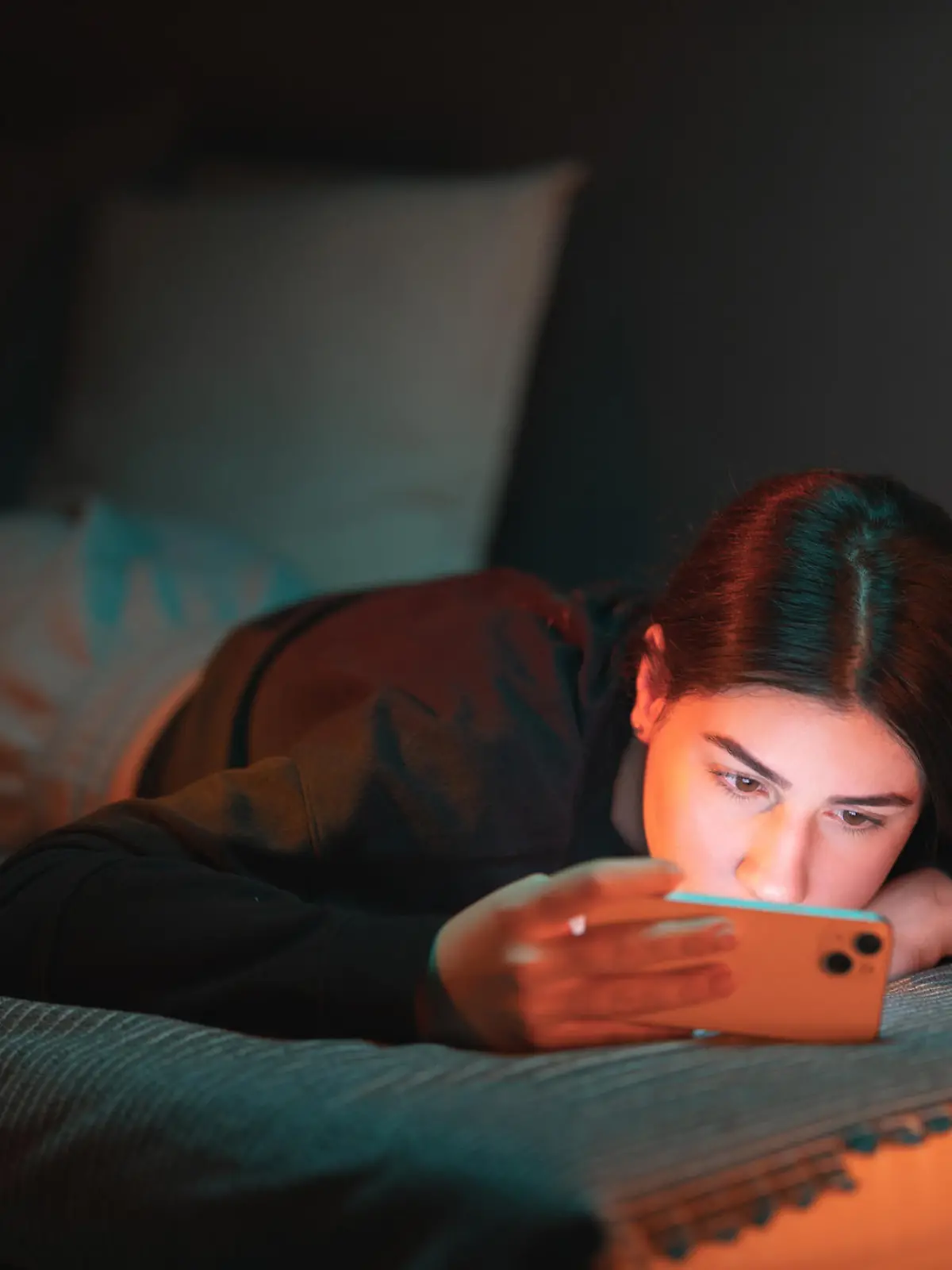 A teenage girl lies on the bed in her room lightened with orange and teal neon lights and watches a movie on her mobile phone.