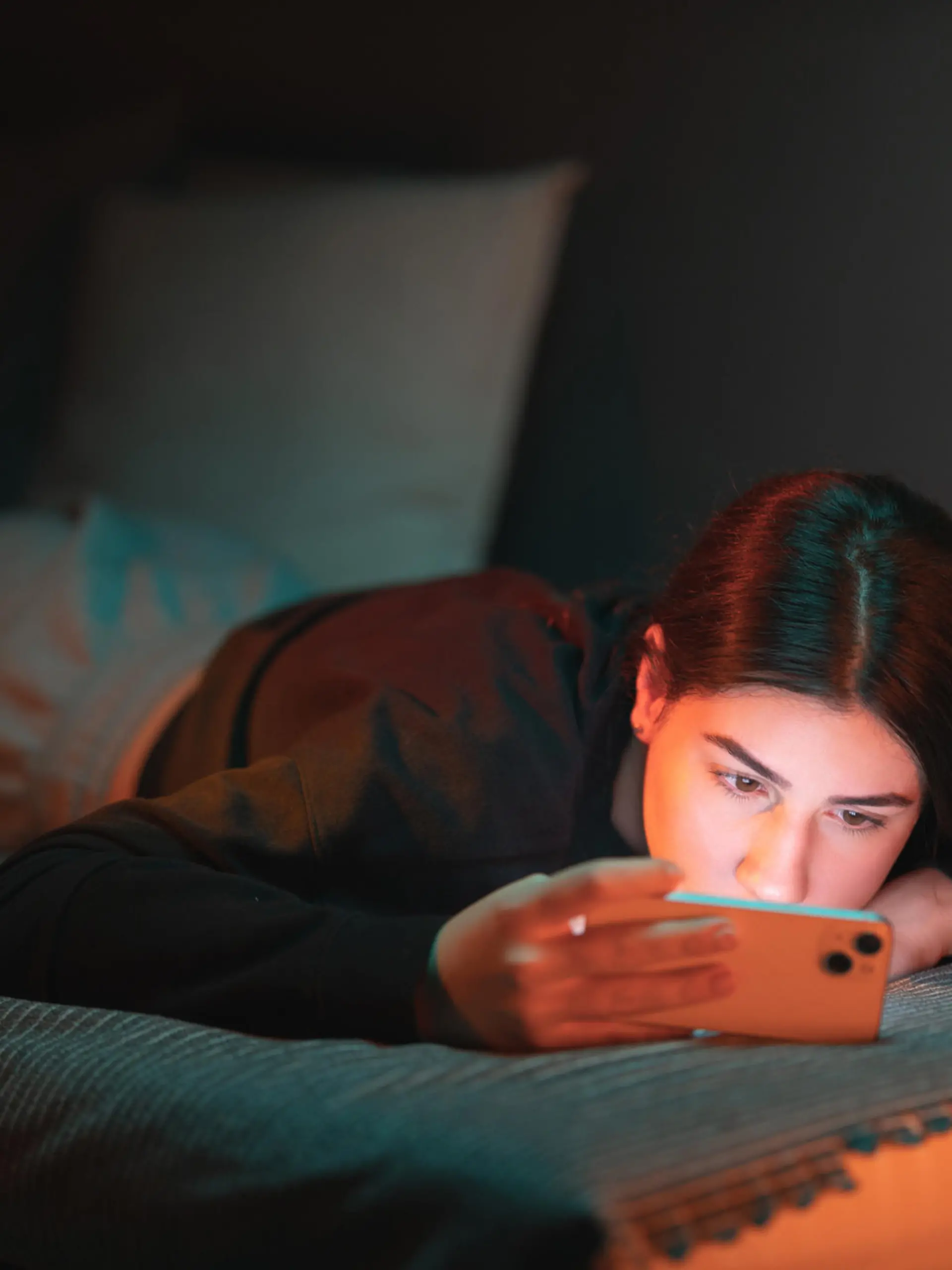 A teenage girl lies on the bed in her room lightened with orange and teal neon lights and watches a movie on her mobile phone.