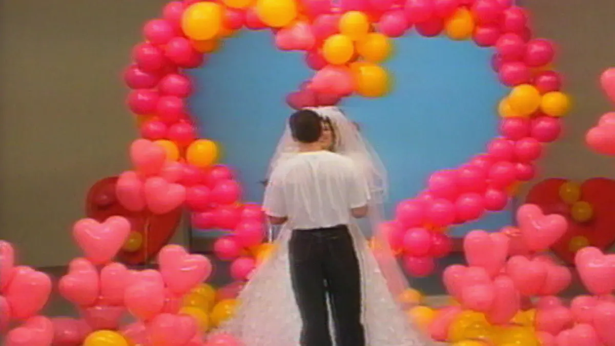 Rene steht vor einem Herz aus Luftballons und bekommt einen Heiratsantrag von seiner Freundin im Brautkleid.