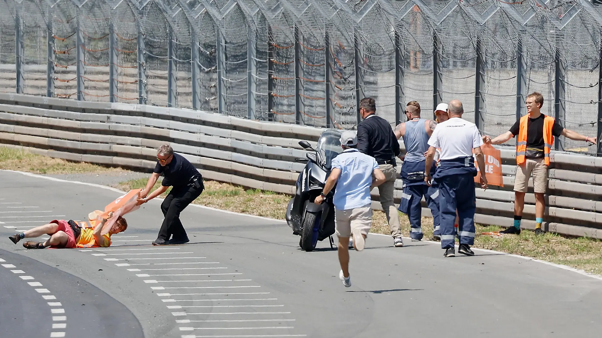 Streckenposten zerren einen Aktivisten der Letzten Generation mit Protestbanner in der Hand von der Fahrbahn, während ein Aktivist am Sicherheitszaun klebt. Am Wochenende findet das 80. Internationale ADAC Norisring Speedweekend statt.