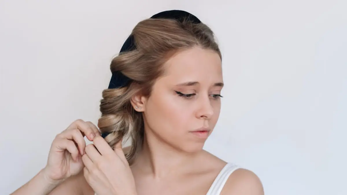 Heatless curl. A young attractive caucasian blonde woman removing soft curler from the hair on a light background. Healthy hair styling, no heating, no damage. A girl twisting curls around the rim