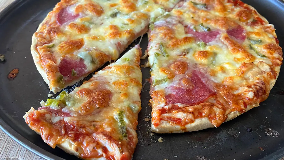 Angeschnittene, aufgebackene Tiefkuehlpizza mit viel Kaese und Salami auf einem Pizzablech,das gerade aus dem Backofen genommen wurde