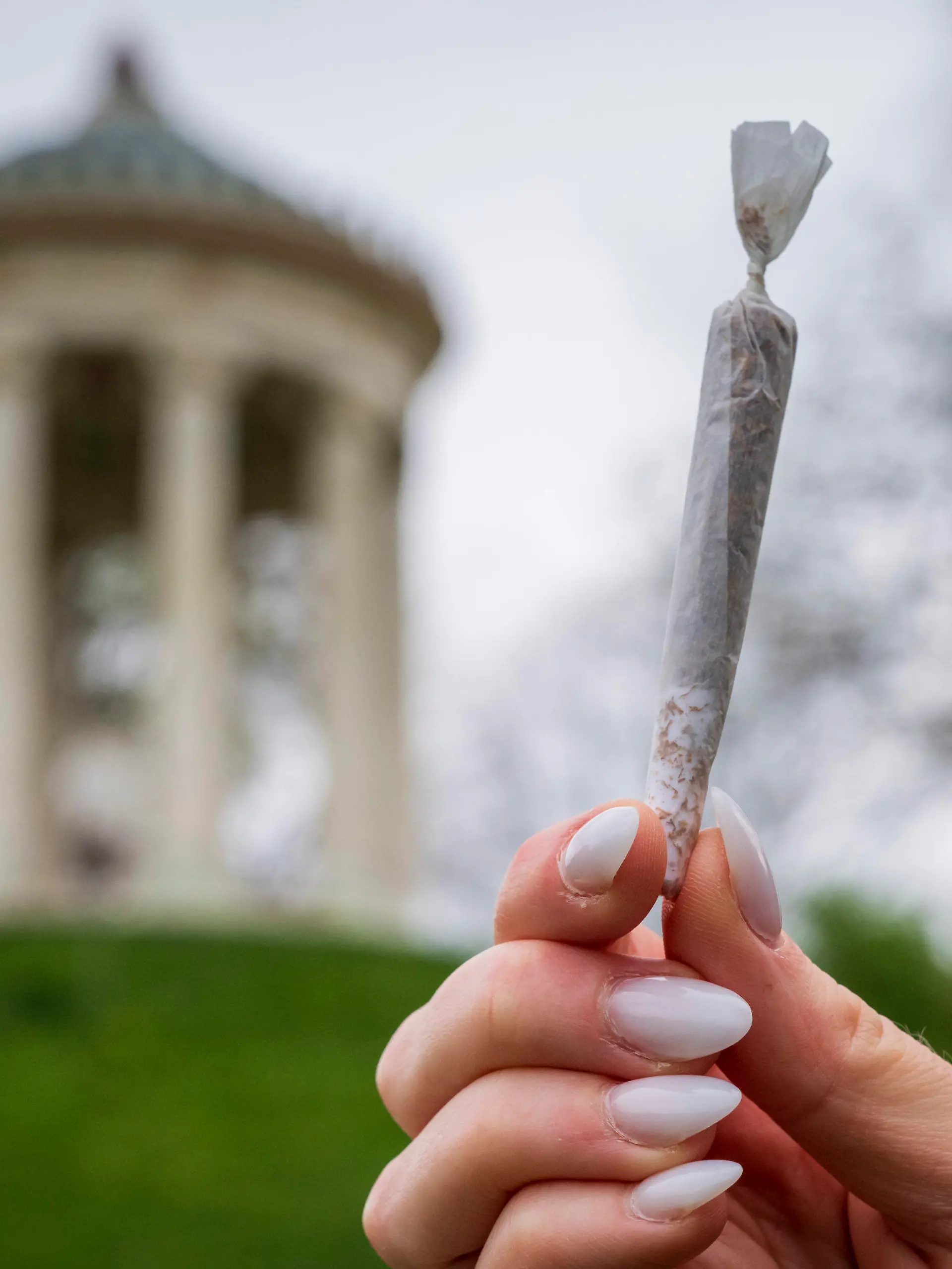 ILLUSTRATION - 10.04.2024, Bayern, München: Eine Frau hält einen Joint vor der Kulisse des Monopteros im Englischen Garten. Seit Ostermontag gelten weite Teile des umstrittenen Cannabisgesetzes. Damit ist fortan für Erwachsene ab 18 Jahren der Besitz von bis zu 25 Gramm Cannabis zum Eigenkonsum erlaubt. In der eigenen Wohnung sollen drei Cannabispflanzen erlaubt sein und bis zu 50 Gramm Cannabis zum Eigenkonsum. Verboten bleibt Kiffen allerdings im öffentlichen Raum unter anderem in Schulen, Sportstätten und in Sichtweite zu solchen Einrichtungen. (zu dpa: «Kiffen im Englischen Garten in München von Donnerstag an verboten») Foto: Peter Kneffel/dpa +++ dpa-Bildfunk +++