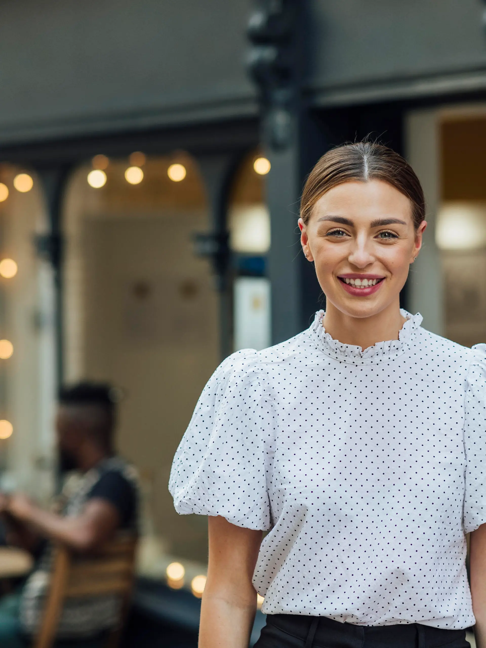 Waist up portrait of a young beautiful woman dressed formally with her hair tied back standing outside of a cafe looking at the camera. There are students out of focus sitting at an outdoor cafe table behind her. She is very cheerful looking at the camera.