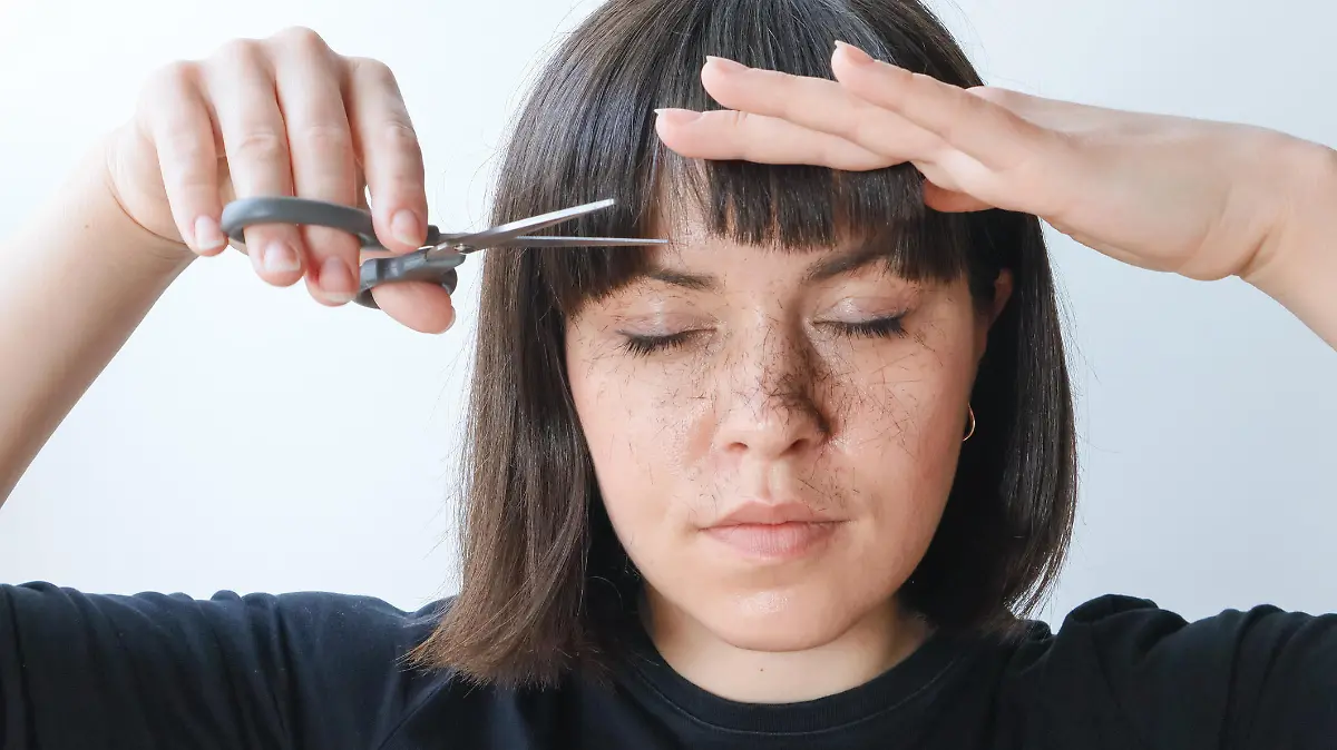 A woman, a brunette girl holds scissors in her hands and cuts her bangs. Hair is cut and falls on the face, portrait.