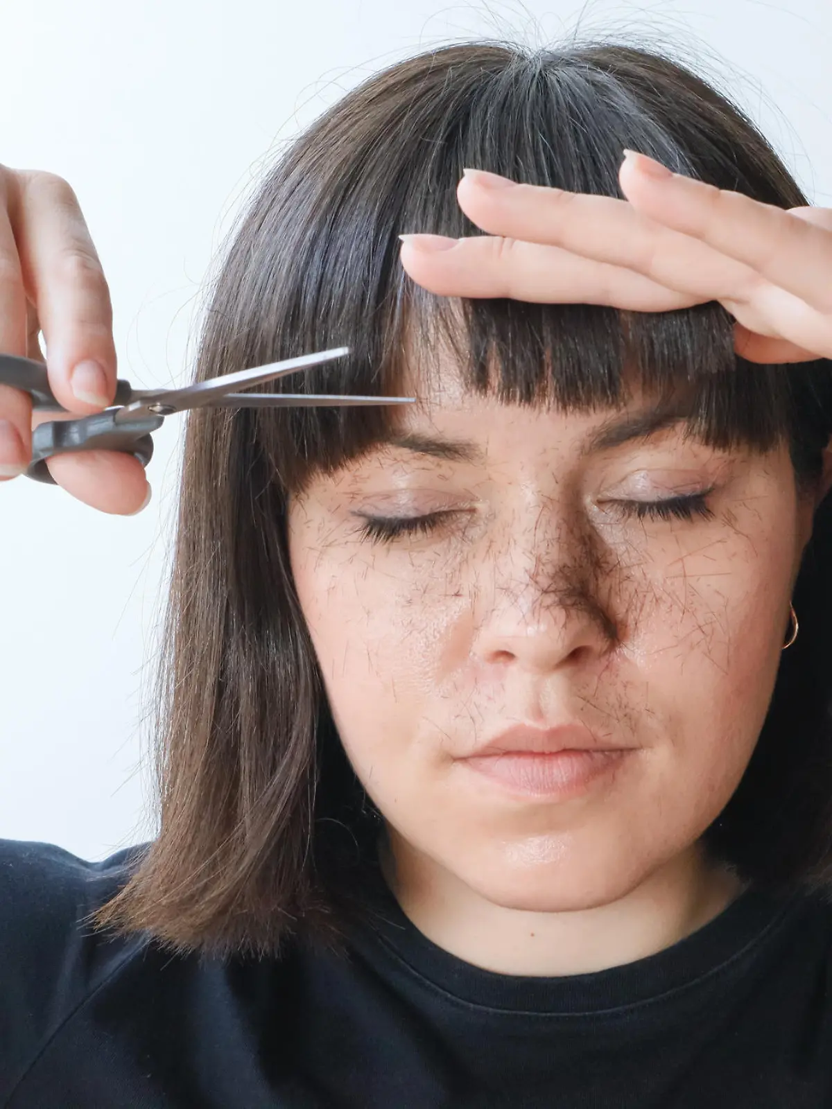 A woman, a brunette girl holds scissors in her hands and cuts her bangs. Hair is cut and falls on the face, portrait.