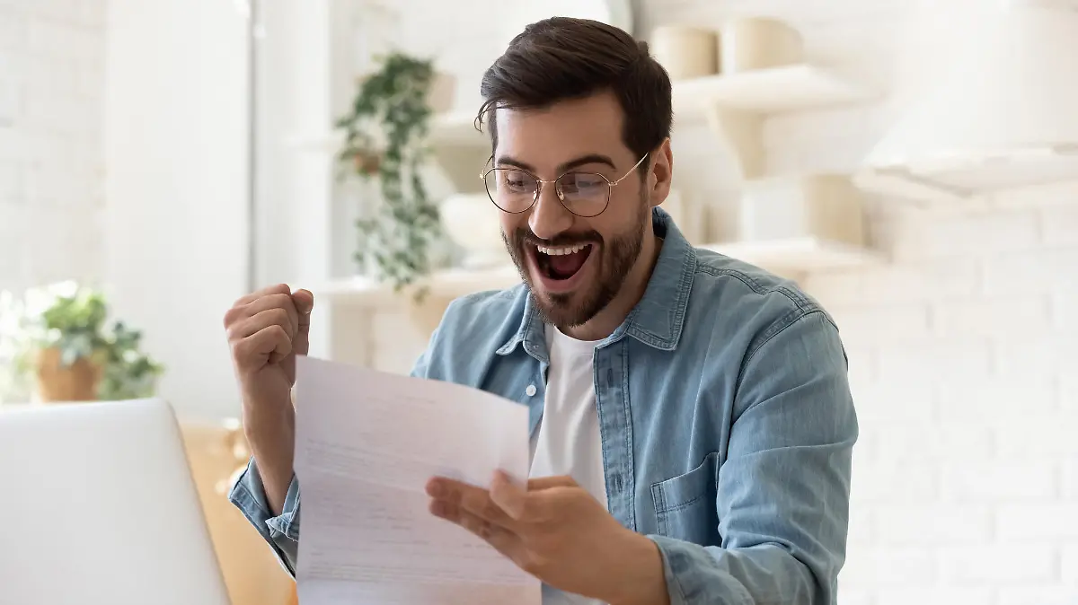 Excited euphoric happy young man holding reading paper postal mail letter amazed overjoyed by good news, got new job celebrate taxes refund receive salary payment loan approval sit at home table