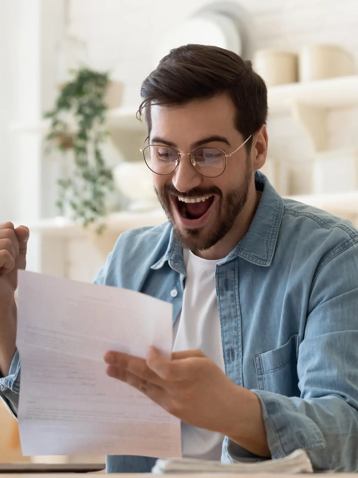 Excited euphoric happy young man holding reading paper postal mail letter amazed overjoyed by good news, got new job celebrate taxes refund receive salary payment loan approval sit at home table