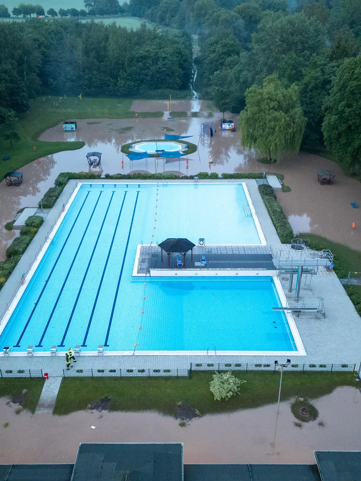25.05.2024 / Bad Berka / Kurz nach 20 Uhr kam es heute in Bad Berka, Neusaalborn und Blankenhain zu einem Unwetter mit Starkregen. Laut Augenzeugen sind innerhalb kürzester Zeit mehr als 30 Liter Regen gefallen. Das Wasser schoss über die Felder von Blankenhain (nähe Golfresort, wo in den kommenden Tagen die Deutsche Nationalmannschaft trainiert) durch Neusaalborn und Bad Berka. In Bad Berka richtete das Hochwasser starke Schäden am Freibad der Stadt an. Der Klingelbach, welcher neben dem Freibad einen Bachlauf hat, trat innerhalb kürzester Zeit massiv über die Ufer und überflutete das Gelände des Freibads. Das Wasser stand in den Gebäuden des Bads mehr als 30 cm hoch. Glücklicherweise wurde die Pumpenanlage nicht beschädigt.
Die Schäden am Freibad sind dennoch sehr hoch. Die Feuerwehr pumpte am Abend mit einer Saugpumpe Wasser ab. Einsatzkräfte entfernten mit Wasserschiebern Wasser aus dem Gebäude.
In den kommenden Tagen muss das Freibad geschlossen bleiben. Dann wird geprüft, wie hoch die Schäden sind.
Auch im Stadtgebiet von Bad Berka sorgte das Wasser für diverse Schäden. Die Trebestraße wurde komplett überflutet. Hier musste die Feuerwehr die Straße sperren und aufwendig beräumen.

Ob es Schäden rund um das Golfresort bei Blankenhain gibt, lässt sich zum Stand heute Abend nicht sagen. Augenzeugen berichteten hier, dass das Wasser sich auch hier seinen Weg suchte.
/ Foto: Johannes Krey | JKFTV.de
