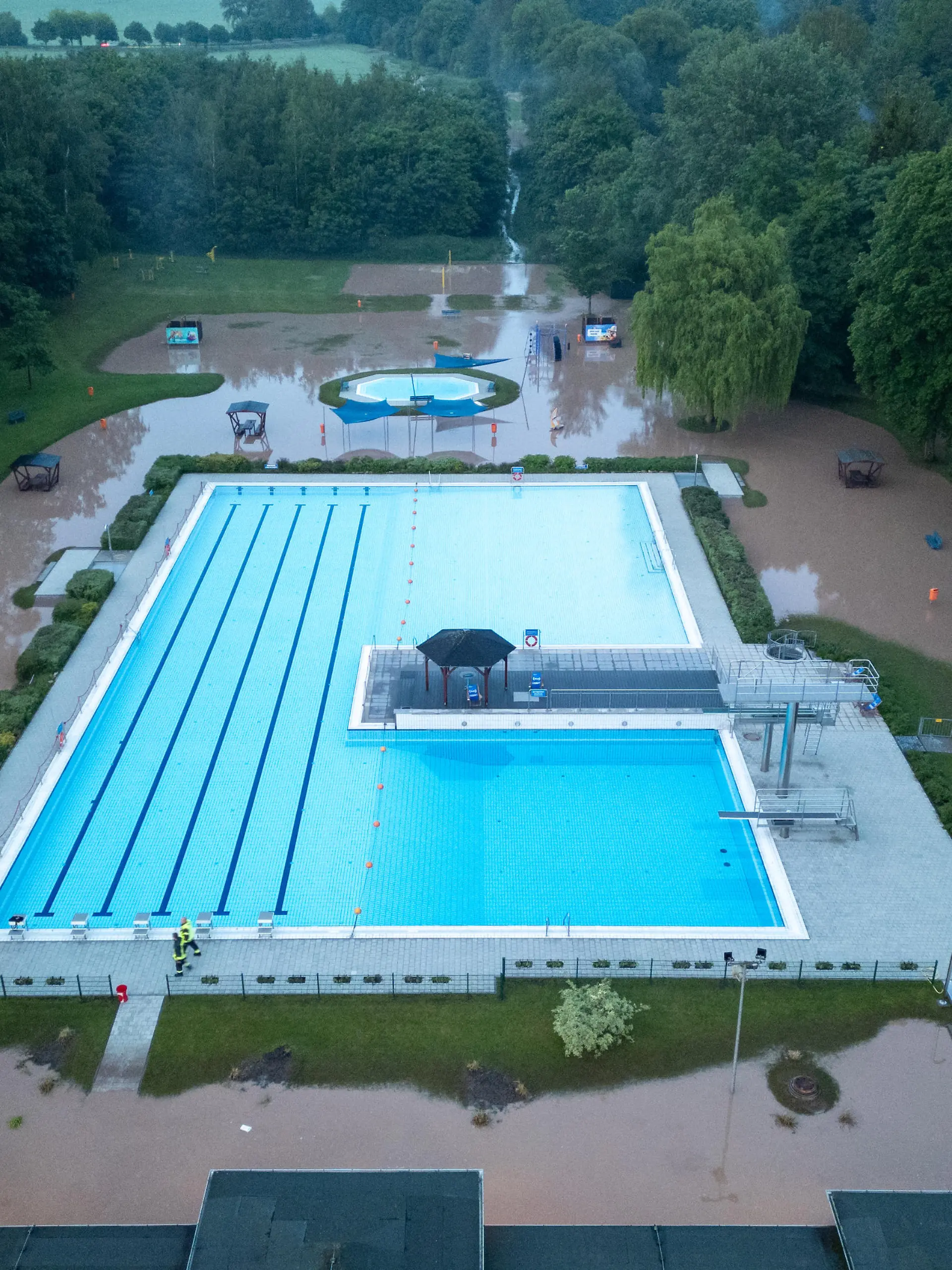 25.05.2024 / Bad Berka / Kurz nach 20 Uhr kam es heute in Bad Berka, Neusaalborn und Blankenhain zu einem Unwetter mit Starkregen. Laut Augenzeugen sind innerhalb kürzester Zeit mehr als 30 Liter Regen gefallen. Das Wasser schoss über die Felder von Blankenhain (nähe Golfresort, wo in den kommenden Tagen die Deutsche Nationalmannschaft trainiert) durch Neusaalborn und Bad Berka. In Bad Berka richtete das Hochwasser starke Schäden am Freibad der Stadt an. Der Klingelbach, welcher neben dem Freibad einen Bachlauf hat, trat innerhalb kürzester Zeit massiv über die Ufer und überflutete das Gelände des Freibads. Das Wasser stand in den Gebäuden des Bads mehr als 30 cm hoch. Glücklicherweise wurde die Pumpenanlage nicht beschädigt.
Die Schäden am Freibad sind dennoch sehr hoch. Die Feuerwehr pumpte am Abend mit einer Saugpumpe Wasser ab. Einsatzkräfte entfernten mit Wasserschiebern Wasser aus dem Gebäude.
In den kommenden Tagen muss das Freibad geschlossen bleiben. Dann wird geprüft, wie hoch die Schäden sind.
Auch im Stadtgebiet von Bad Berka sorgte das Wasser für diverse Schäden. Die Trebestraße wurde komplett überflutet. Hier musste die Feuerwehr die Straße sperren und aufwendig beräumen.

Ob es Schäden rund um das Golfresort bei Blankenhain gibt, lässt sich zum Stand heute Abend nicht sagen. Augenzeugen berichteten hier, dass das Wasser sich auch hier seinen Weg suchte.
/ Foto: Johannes Krey | JKFTV.de