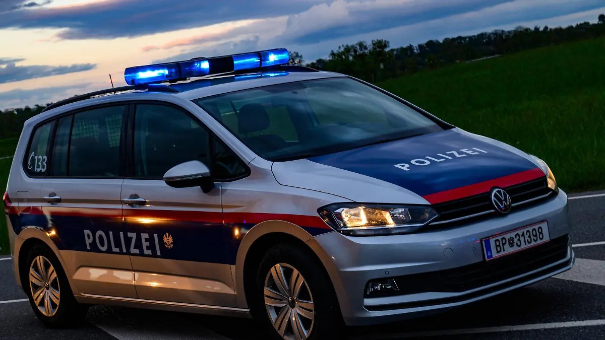 Ein Polizeifahrzeug der österreichischen Polizei steht einsatzbereit an der Straßenseite. Die Dämmerung setzt ein, während die Blaulichter des Fahrzeugs hell in den frühen Abendhimmel strahlen und die Umgebung in ein weiches Licht tauchen. - 20240421_PD18121