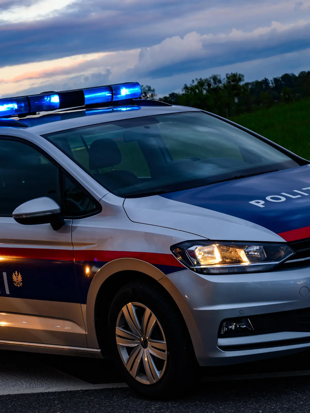 Ein Polizeifahrzeug der österreichischen Polizei steht einsatzbereit an der Straßenseite. Die Dämmerung setzt ein, während die Blaulichter des Fahrzeugs hell in den frühen Abendhimmel strahlen und die Umgebung in ein weiches Licht tauchen. - 20240421_PD18121