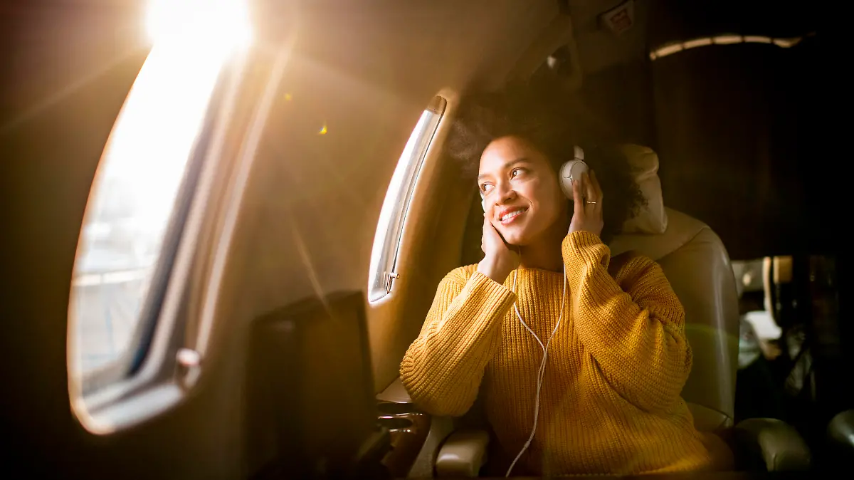 Young fashionable woman sitting on a private airplane and looking through a window while listening to music through headphones.