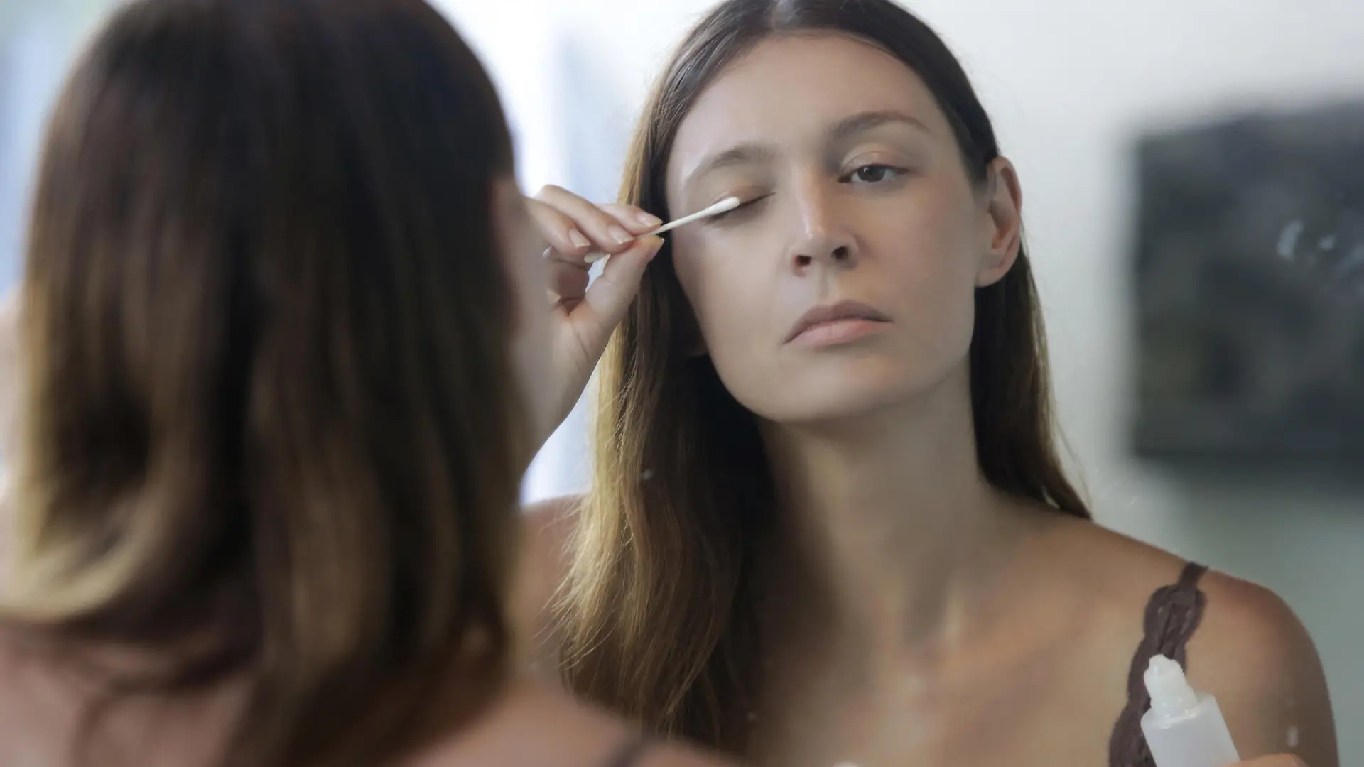 Young caucasian woman applying serum for eyebrows and eyelashes growth in front of the mirror. Night time beauty routine.