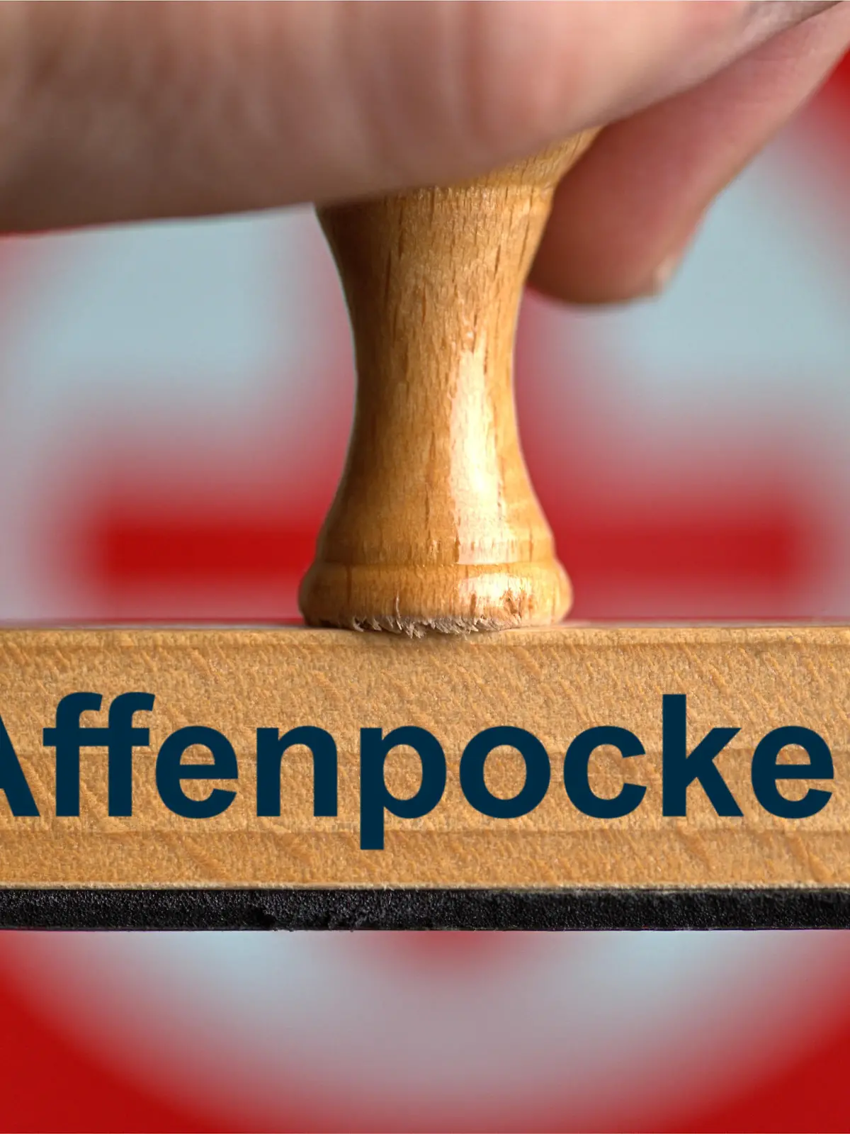 [M] Ein symbolischer Holzstempel mit der Aufschrift "Affenpocken", gehalten von einer Hand im Anschnitt vor einem unscharfen Hintergrund mit einem roten Kreuz. Foto mit Composing als Symbolbild für das vermehrte Auftreten der Affenpocken bei Menschen. || Modellfreigabe vorhanden
