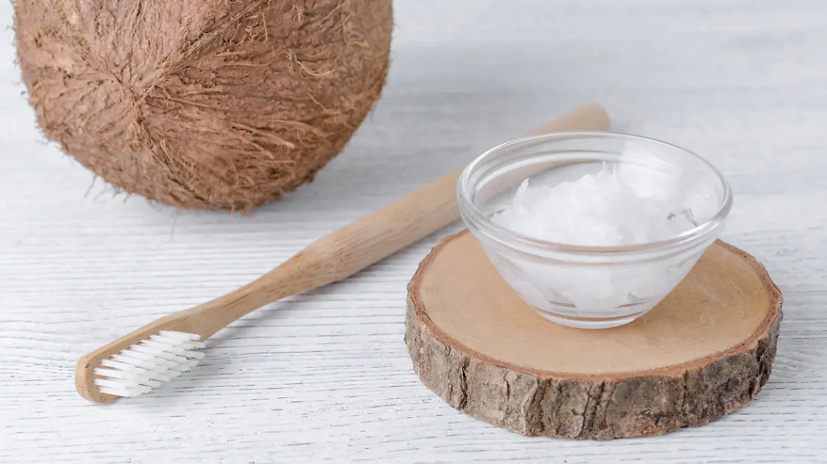 Eine Kokosnuss, eine Holzzahnbürste und ein Schälchen mit Kokosöl in einer kleinen durchsichtigen Schale auf einem weißen Holzuntergrund.