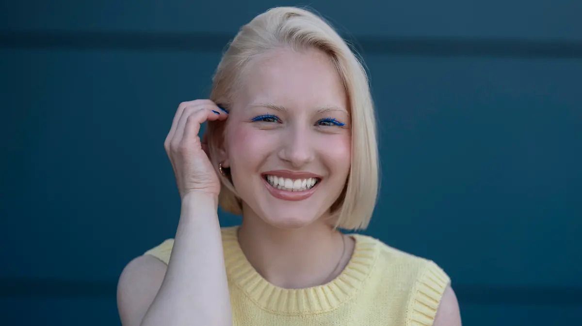 A beautiful blonde girl with chin-length hair, with blue mascara looks at the camera and smiles with teeth, behind her is a dark blue background.