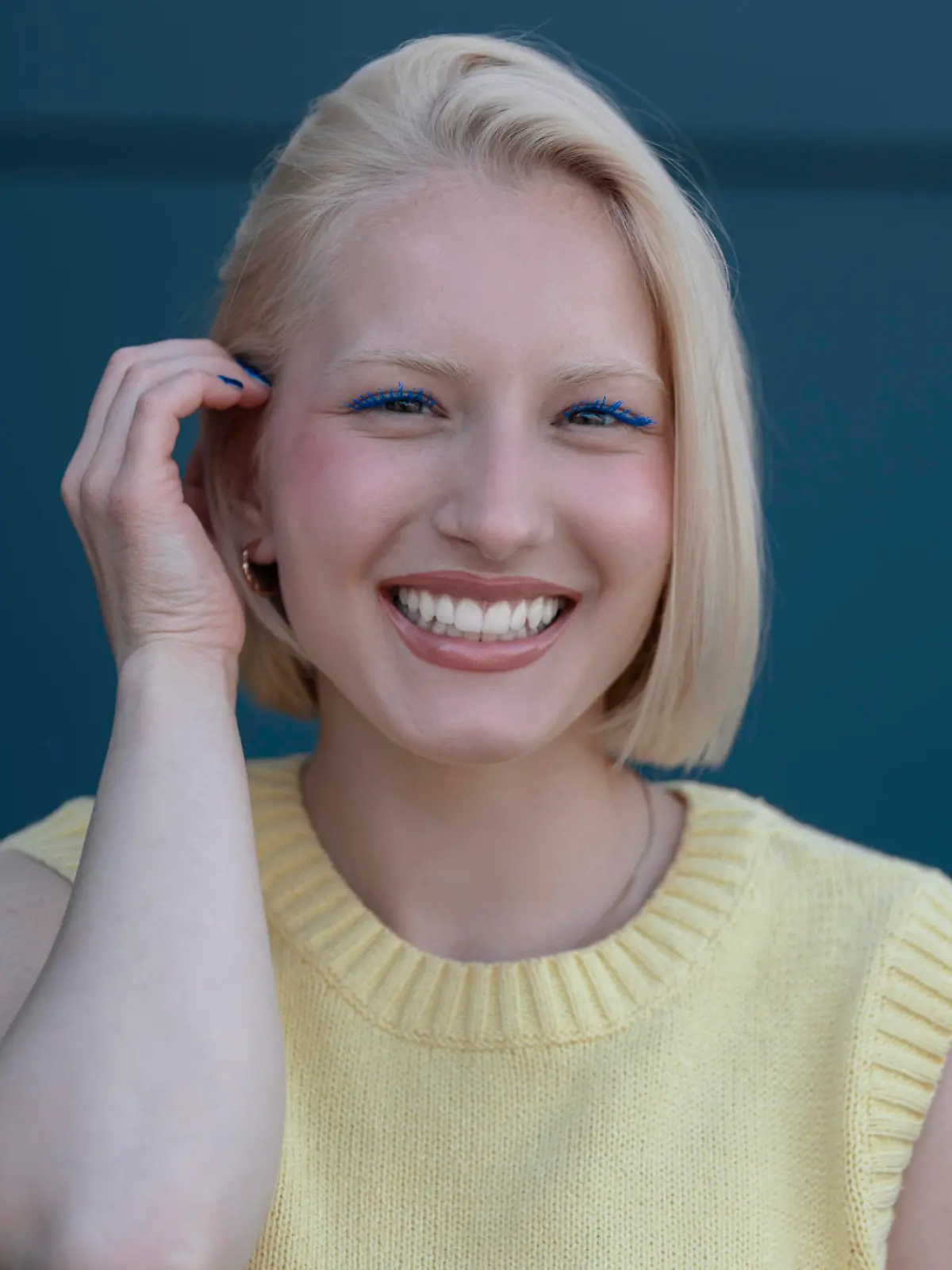 A beautiful blonde girl with chin-length hair, with blue mascara looks at the camera and smiles with teeth, behind her is a dark blue background.