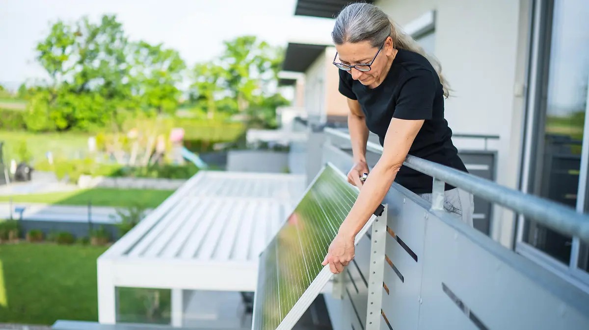 mature adult woman wearing glasses installing adjustable solar panel on balcony of her house in estate of terraced houses in rural landscape on sunny morning