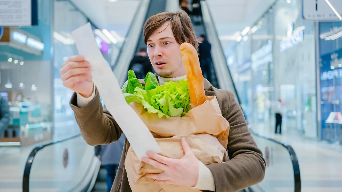 Man in a coat holds a paper check and a bag with a baguette and lettuce in a shopping mall and is shocked by the high prices of groceries.