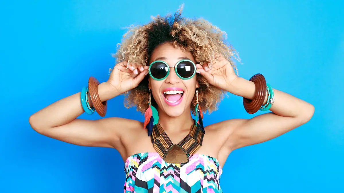 Summer portrait of happy afro american young woman wearing summer clothes and sunglesses, standing against blue background and laughing at camera.