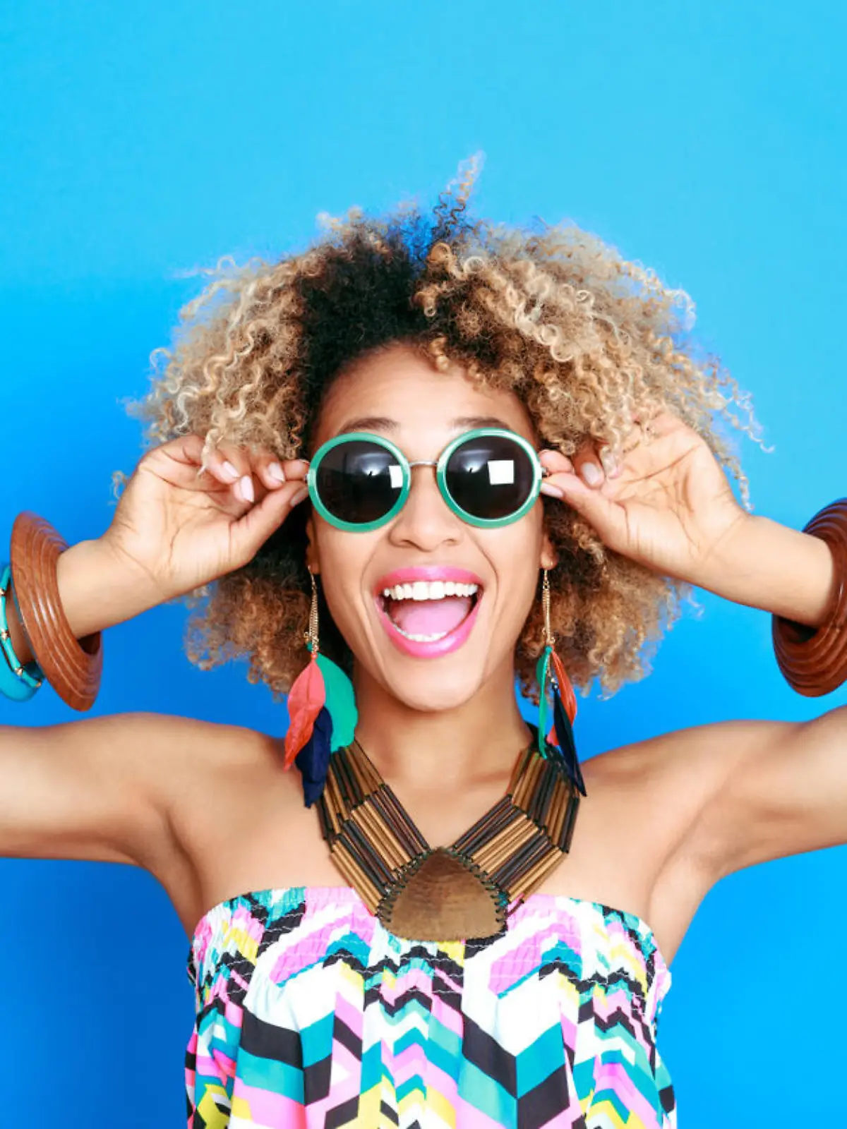 Summer portrait of happy afro american young woman wearing summer clothes and sunglesses, standing against blue background and laughing at camera.