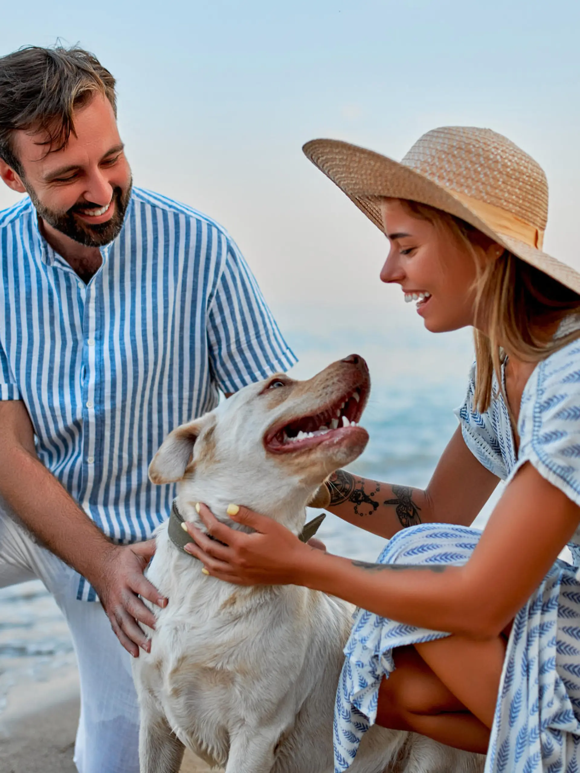 A cute woman in a dress and a straw hat and a handsome man in a striped shirt with their labrador dog are having fun on the seashore.