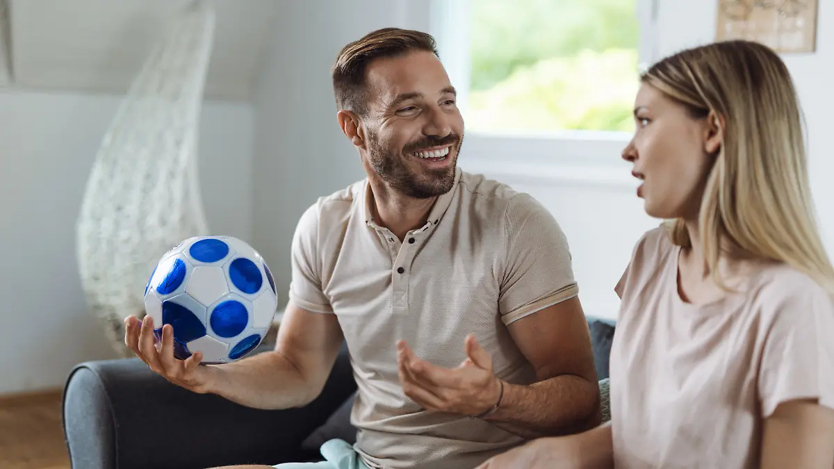 Ein Paar sitzt auf der Couch und schaut Fußball. Die Partnerin schaut genervt. 